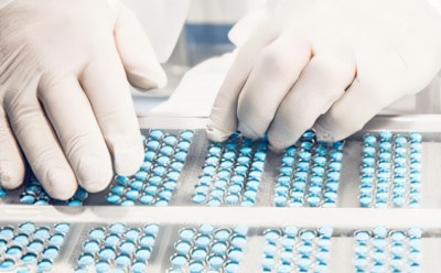 A pharmaceutical or medical manufacturing facility. It shows a person in protective clothing handling trays filled with blue capsules.