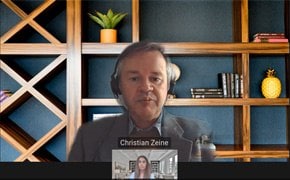 A virtual meeting screenshot with Christian Zane and another participant. A small window shows the other participant speaking. In the background, a well-organized shelf holds books, decorative objects, and a potted plant.