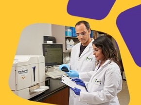 Two lab technicians in white lab coats are collaborating at a laboratory bench, one holding a clipboard and the other using a device. The background features various laboratory supplies and equipment, with a vibrant yellow and purple design framing the image.