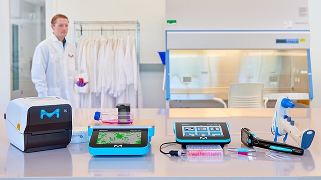 Five instruments sitting on a laboratory bench with a scientist in a lab coat walking past in the background. Five instruments sitting on a laboratory bench with a scientist in a lab coat walking past in the background.