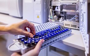 A close-up image of a person's hand inserting a blue and black sample tray into a laboratory instrument. The background shows part of the machine with visible components, emphasizing a laboratory setting.