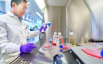Scientist at the Tissue Culture Hood Male scientist sitting at the tissue culture hood holding a handheld pipette gun pipetting red media into a six well cell culture plate. Three media bottles are in the background and a T flask with media is in the foreground on a Millicell® DCI.