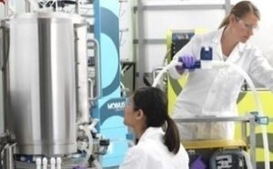 Two scientists work to the right of a bioreactor