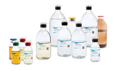 A collection of laboratory bottles containing various sterility testing culture media rinse and fluid. There are large transparent bottles with blue labels in the back row, medium-sized transparent and amber-colored bottles with different colored caps in the middle, and small transparent bottles with red, green, yellow, and blue caps in the front row. Each bottle has a label indicating its contents or purpose within sterility testing procedures.