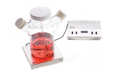 A laboratory setup featuring an induction stirrer with a red liquid in a glass container placed on it, showcasing two control knobs and an on/off switch on a white base, against a plain background.