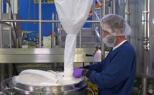 In the image, a person in a blue gown and hairnet is pouring a white substance from a large bag into an industrial mixing bowl in a manufacturing facility.