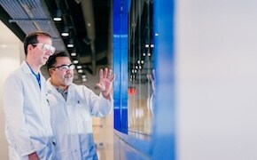 Two individuals in white lab coats discussing in front of a large screen in a modern lab.