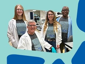 A group of four individuals in a laboratory setting, all wearing matching gray t-shirts with a blue design on the front. They are dressed in white lab coats, and one person is seated while the others stand beside them. The background features laboratory equipment, suggesting an environment focused on scientific work.