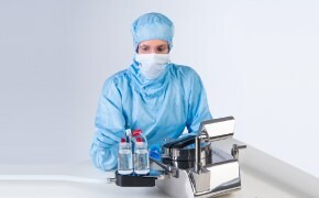 Öt fehér laborköpenyt, védőszemüveget és kék nitrilkesztyűt viselő tudós dolgozik a laboratórium különböző állomásain, ahol kémiai analitikai műszereket kezelnek, és üvegpoharakat és üvegcséket töltenek. 