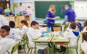 몇 명의 학생들이 실험용 가운과 장갑을 착용하고 책상에 앉아 있습니다. 머크의 자원봉사자 두 명이 돌아다니며 학생들을 도와줍니다.