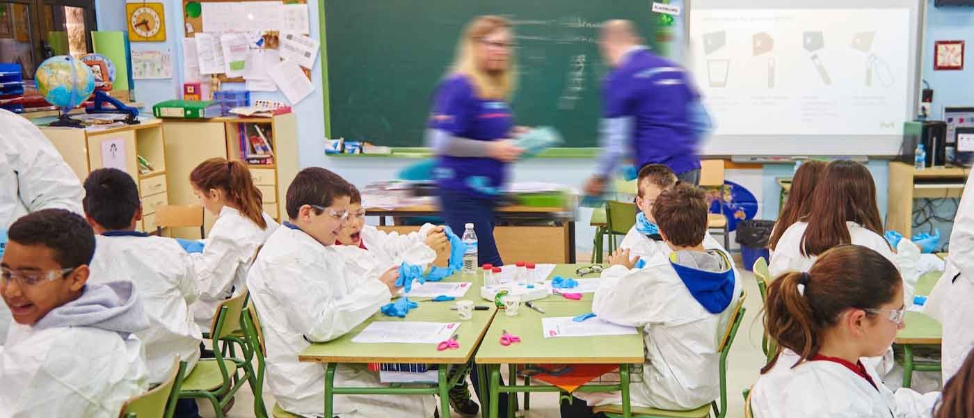 Curiosity Lab Classroom Scene 학생들은 실험용 가운과 장갑을 착용하고 책상에 앉아 있습니다. 당사의 자원봉사자 두 명이 돌아다니며 학생들을 도와줍니다.