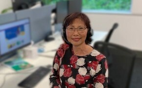 Grace Yee는 헤드셋을 착용하고 책상에 앉아 있습니다. 뒤에는 데스크톱 컴퓨터가 있습니다.