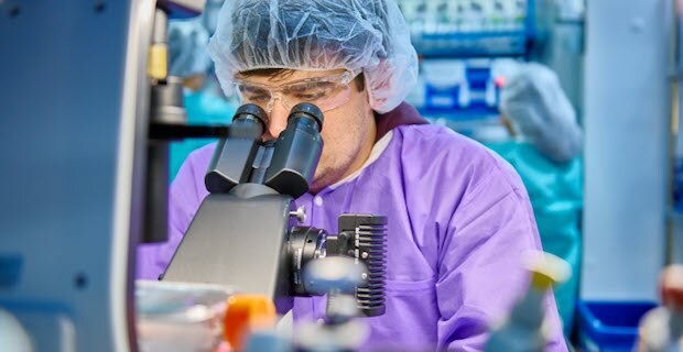 Innovative products expertise - Scientist using microscope 연구자가 보라색 실험복, 보안경, 머리망을 착용하고 현미경을 들여다보고 있습니다.