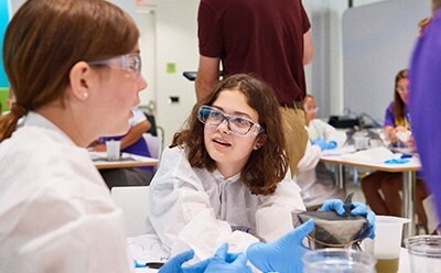 Two children are engaged in a hands-on science activity, wearing protective lab coats and gloves. One child, holding a container, is demonstrating something to another child who is attentively listening, while a few adults and other participants are visible in the background, creating an interactive and educational environment.